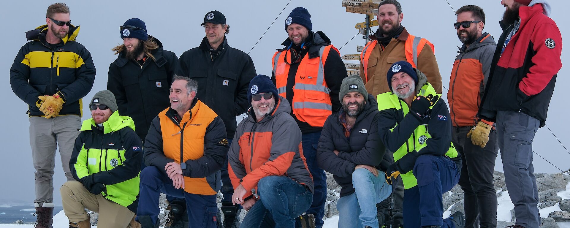 12 Expeditioners pose for a photo on the snow in front of the Casey station sign