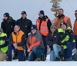 12 Expeditioners pose for a photo on the snow in front of the Casey station sign