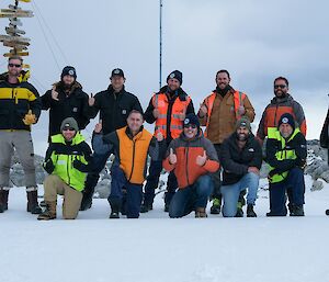 12 Expeditioners pose for a photo on the snow in front of the Casey station sign