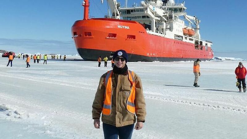 A man in a brown jacket stand on ice in front of RSV Nuyina.