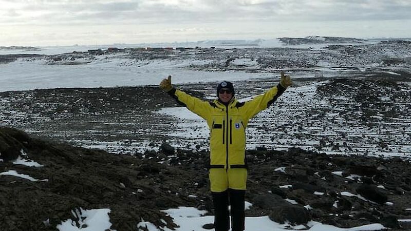A man in yellow expeditioner garb stands with his arms up on rocks and ice
