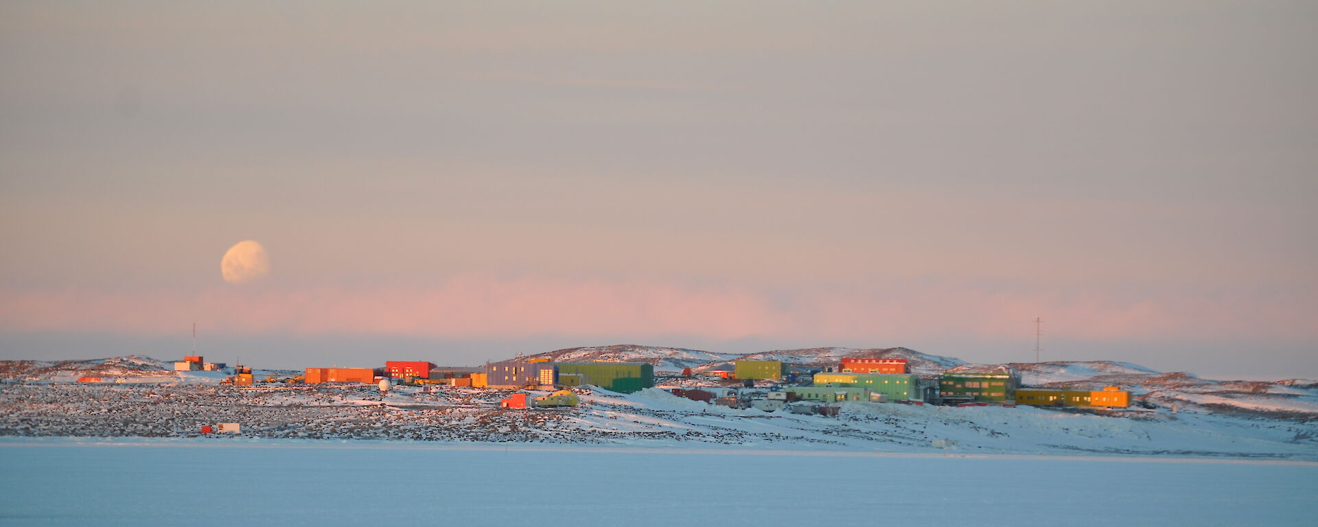 Distant view of colourful buildings across sea ice with a full moon behind.