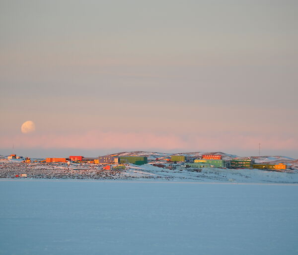Distant view of colourful buildings across sea ice with a full moon behind.