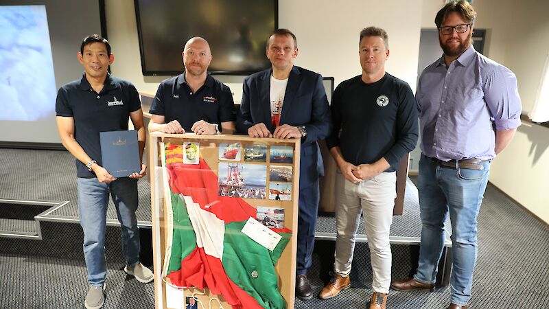 Five men stand behind a glass-framed case with a flag, coin and photos inside.