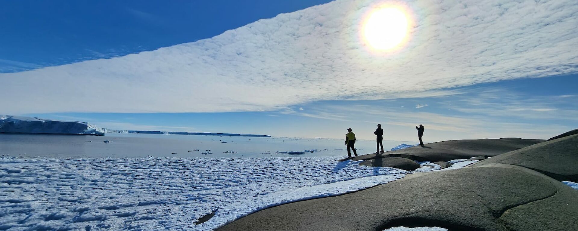 Silhouettes of three expeditioners in the distance as the sun shines through a band of high level cloud