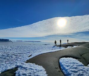 Silhouettes of three expeditioners in the distance as the sun shines through a band of high level cloud