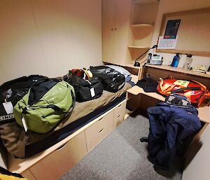 Bags cover a bed made of pale wood, with a desk and cupboards on the right.