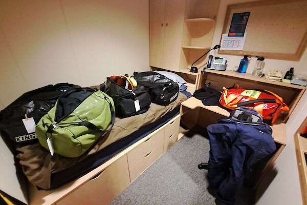 Bags cover a bed made of pale wood, with a desk and cupboards on the right.