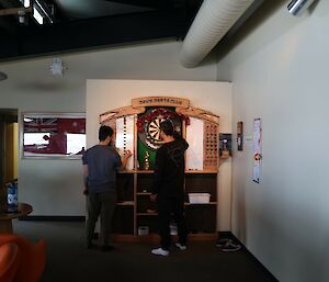 Two men stand in front of a darts board in a white room with a red couch.