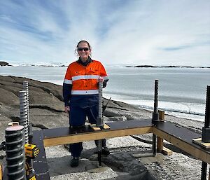 A woman is smiling as she stands next to a bollard foundation on a rocky hillside. In the background is a frozen sea