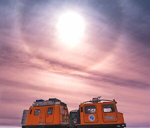 A halo is visible around the sun above two Hägglunds vehicle that are parked on solid ice