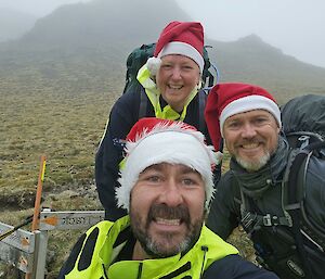 Two men and a woman standing on a foggy hillside with santa hats on