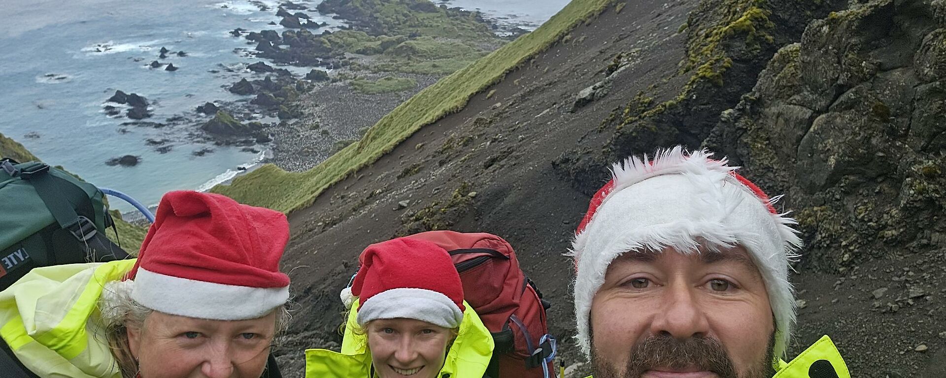 Two men and a woman on a hill with the sea behind them, with santa hats on.