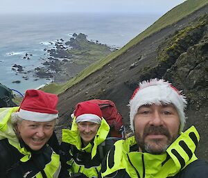 Two men and a woman on a hill with the sea behind them, with santa hats on.