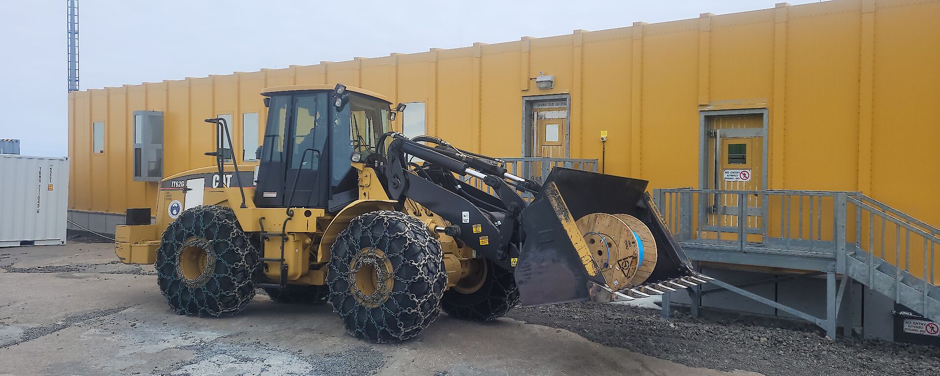 A large roll of fibre optic cable is in the bucket of a loader which is parked outside a yellow building