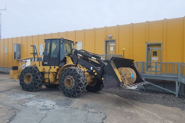 A large roll of fibre optic cable is in the bucket of a loader which is parked outside a yellow building