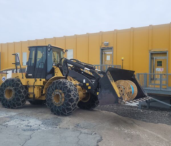 A large roll of fibre optic cable is in the bucket of a loader which is parked outside a yellow building