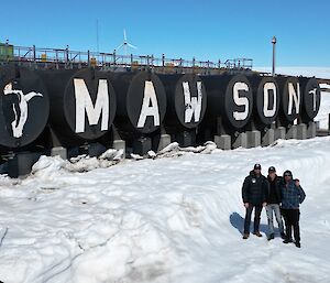 Three men are standing on the snow in front of 8 large fuel tanks with the word 'Mawson' written on them