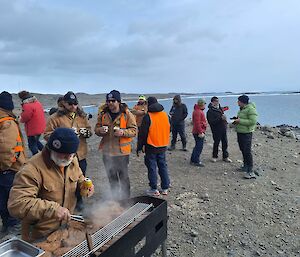 A group of people gather around the shore at Club Lake while one person cooks on a BBQ