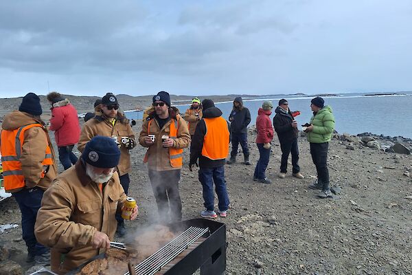 A group of people gather around the shore at Club Lake while one person cooks on a BBQ