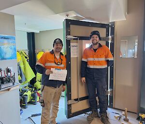 Two men smiling standing in front of an open door holding a sign