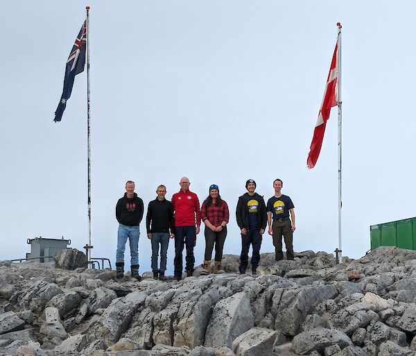 Six people standing between Australian and Danish flags on a rocky outcrop in Antarctica.