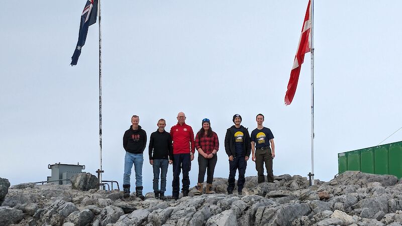 Six people standing between Australian and Danish flags on a rocky outcrop in Antarctica.