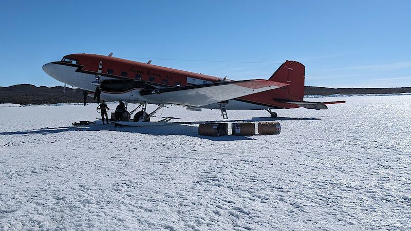 A red and white twin propellor aircraft on skis, parked on ice, with three fuel drums lying beside it.
