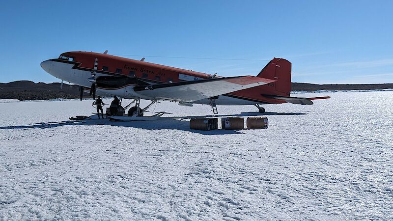 A red and white twin propeller aircraft on skis, parked on ice, with three fuel drums lying beside it.
