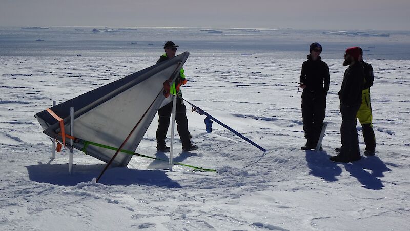 Three people standing beside a reflective triangle, tied down in snow, being prepared for installation on an aircraft.