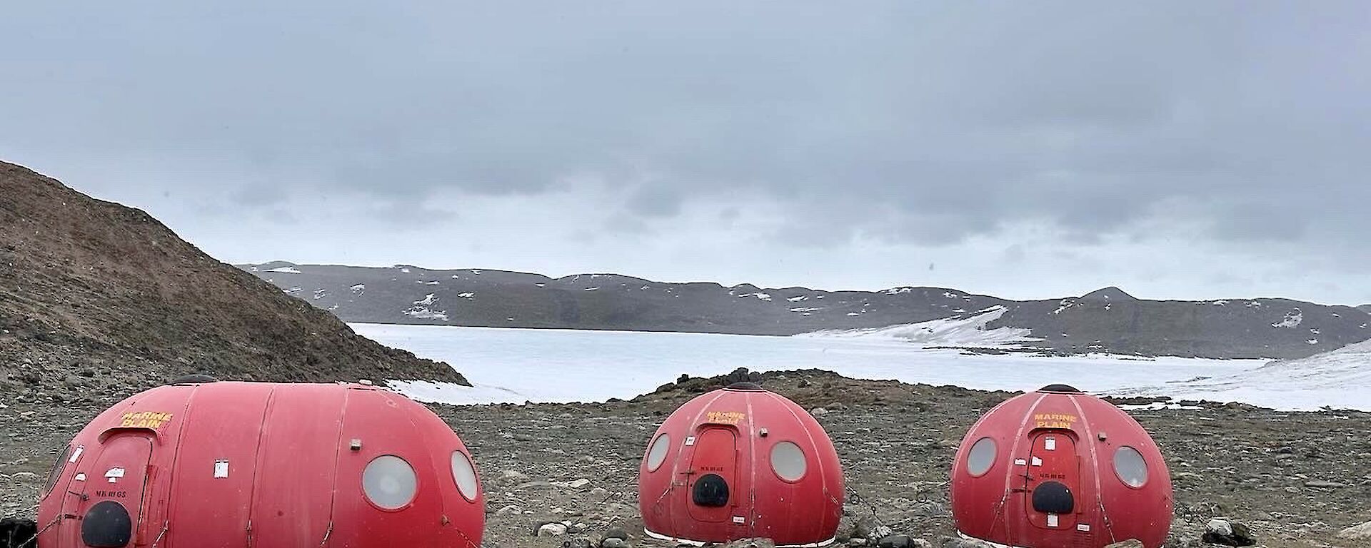 Two round red fibreglass huts and an oblong one sit on a rocky plain with snow in the background