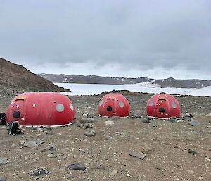 Two round red fibreglass huts and an oblong one sit on a rocky plain with snow in the background
