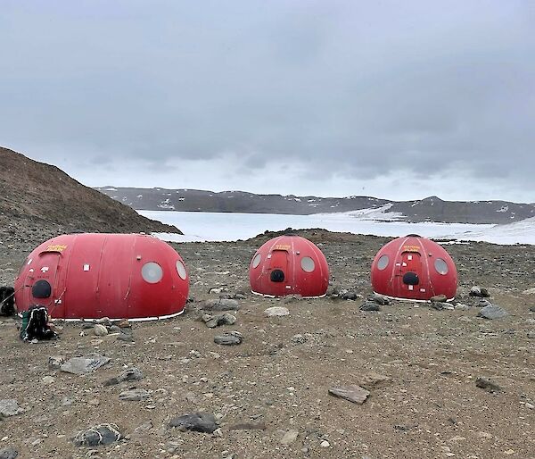 Two round red fibreglass huts and an oblong one sit on a rocky plain with snow in the background