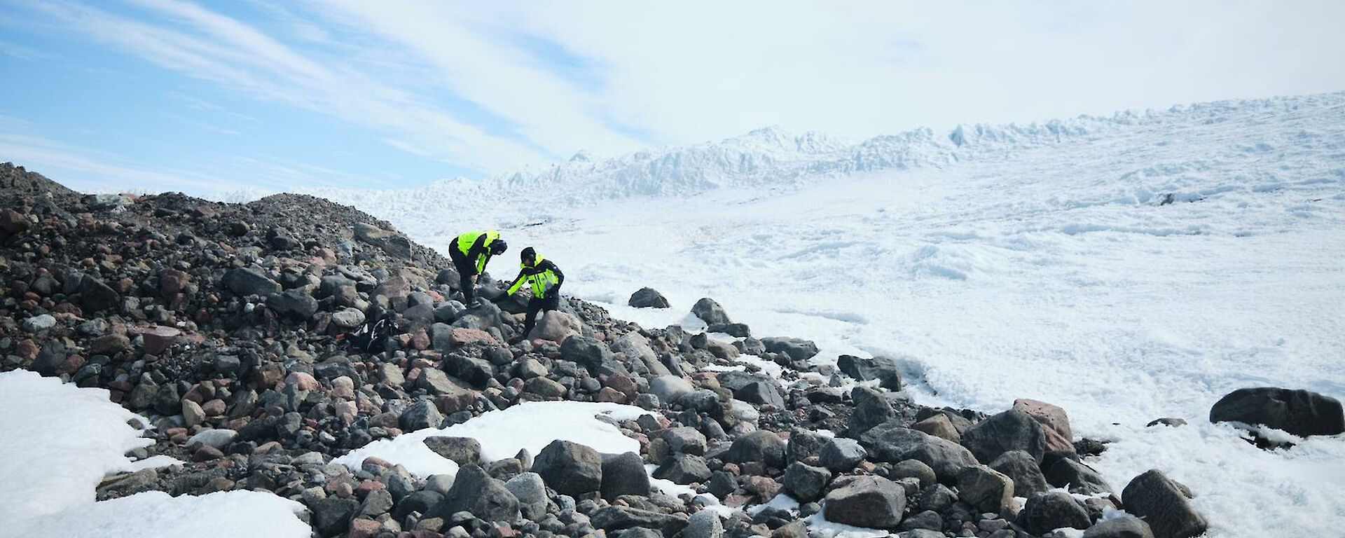 Two people in bright yellow outdoor gear stand on a rocky hillside with snow all around.