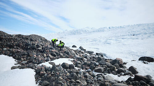Two people in bright yellow outdoor gear stand on a rocky hillside with snow all around.