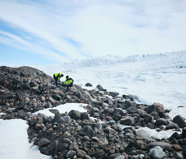 Two people in bright yellow outdoor gear stand on a rocky hillside with snow all around.