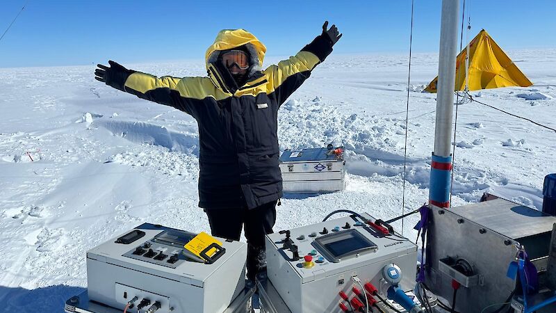 A figure in polar gear stands near equipment on a snowy plain, with arms outstretched