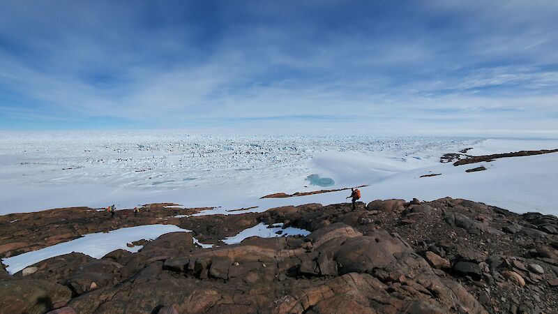 two lone figures on a rocky hillside with a glacier spreading out behind them