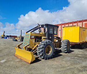 Large yellow vehicle with tractor tyres and digging blade on front with black enclosed cab. looks similar to an old style tractor. Attached is a single axle trailer with a mid length cargo container. In distant background is a crane with cargo containers on ground.