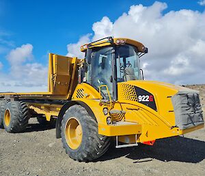 Large 6 wheeled tractor tyred vehicle with a roll off flatbed cargo tray enclosed cab with blue sky and cloud background.