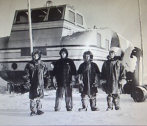 Black and white photo of Large Wheeled vehicle with high enclosed cab depicted front on with 4 figures in parkas and snow clothes in foreground.