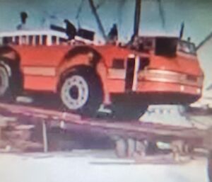 Out of focus shot of Large red wheeled vehicle being offloaded from a ship onto the ice in Antartica. Lone figure in bottom right foreground