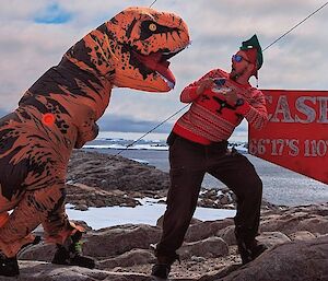one expeditioner dressed in dinosaur costume scares another expeditioner with his tea by the Casey station sign post