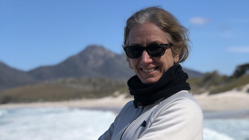 A woman in sunglasses stands with arms crossed, smiling, in front of surf beach