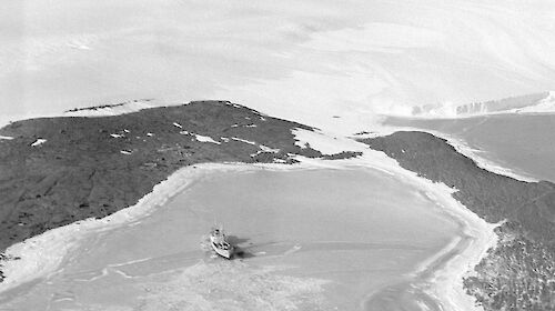 An aerial view of a ship in an ice-filled harbour surrounded by dark rock, with a snow-covered hill rising behind and mountains in the distance.