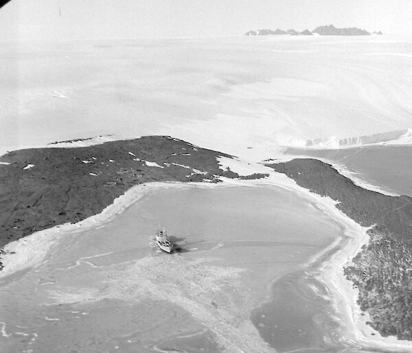 An aerial view of a ship in an ice-filled harbour surrounded by dark rock, with a snow-covered hill rising behind and mountains in the distance.