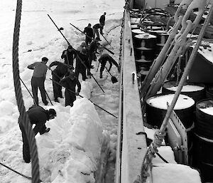 A group of men using crowbars to dig sea ice away from the side of a ship.