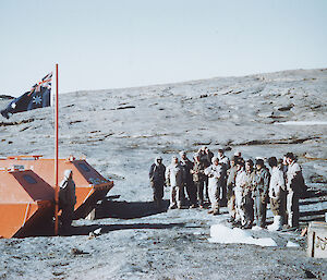 A small group of men stand facing the Australian flag, flying between two small barges. Another man stands by the flagpole.