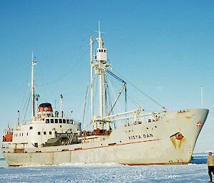 A grey ship parked in sea ice in Antarctica. Two men are walking past its bow.