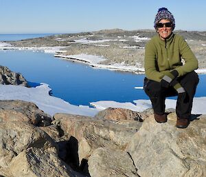 A woman squatting on a rock, with a view of snow and water behind.